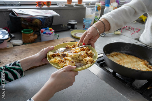 Two people share a plate of freshly cooked crepes at a kitchen counter with various cooking items and utensils around. photo