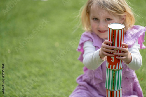 A young girl with blond hair, sitting on grass, is focusing intently on balancing colorful cylindrical blocks in her hands. photo