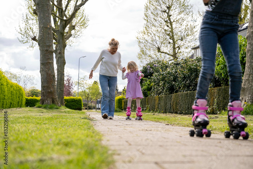 A child in a pink dress learns to roller skate with the help of an adult on a sidewalk lined with greenery under a cloudy sky. photo