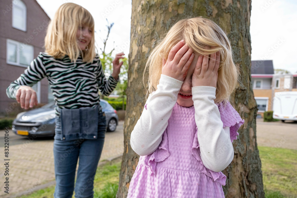 Two children are playing outside near a tree; one child is covering their eyes while the other stands nearby with a playful smile. Two children are playing outside near a tree; one child is covering their eyes while the other stands nearby with a playful smile.