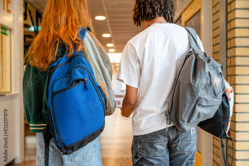 Two students walk down a school hallway, carrying backpacks and apparently engaged in a conversation.