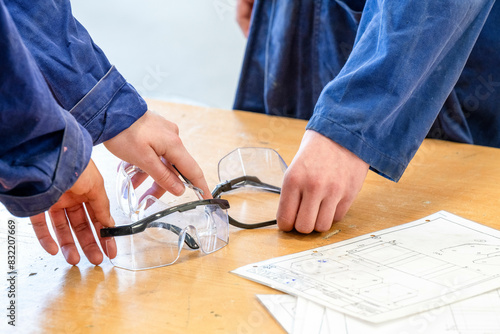 Two pairs of safety glasses rest on a worktable beside technical drawings, with a person's forearms visible, wearing a blue shirt.