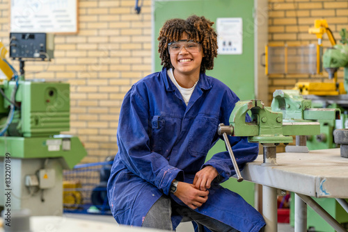 A smiling young person in a blue work overalls sits on a workbench in a workshop with machinery in the background. photo