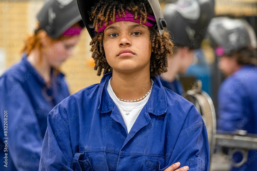 A confident teenager wearing a welding helmet rests arms crossed in a workshop setting, surrounded by peers also dressed in safety gear. A confident teenager wearing a welding helmet rests arms crossed in a workshop setting, surrounded by peers also dressed in safety gear.