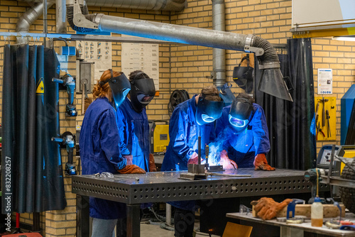Three individuals are engaged in welding activity in a workshop setting, fully equipped with safety gear including helmets and gloves, as sparks fly from the welding point.