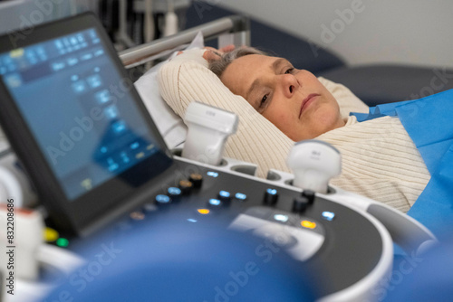 A patient is undergoing an ultrasound exam, with a sonography machine in the foreground and the individual lying down on an examination table wearing a hospital gown. photo
