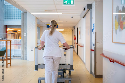 A healthcare professional is walking down a hospital corridor pushing a medical cart, seen from behind. photo