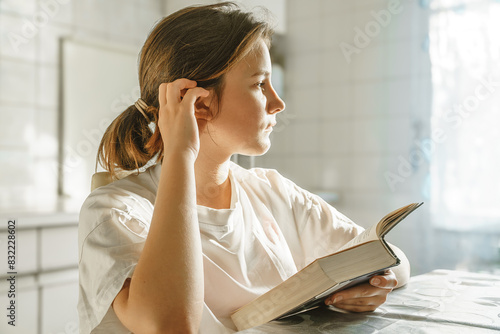 Little cute girl reading a book sitting at a table near the window.