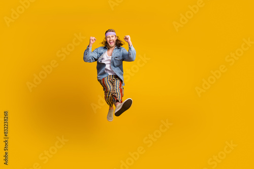 Full size photo of cool young man raise fists empty space jump wear denim shirt isolated on yellow color background