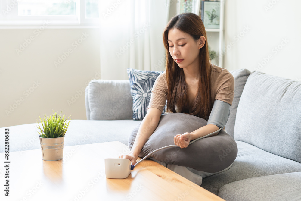 Hypotension, asian young woman sitting on couch checking blood high pressure and heart rate with digital pressure monitor machine making self check up on her arm at home. Healthcare and medical. Hypotension, asian young woman sitting on couch checking blood high pressure and heart rate with digital pressure monitor machine making self check up on her arm at home. Healthcare and medical.