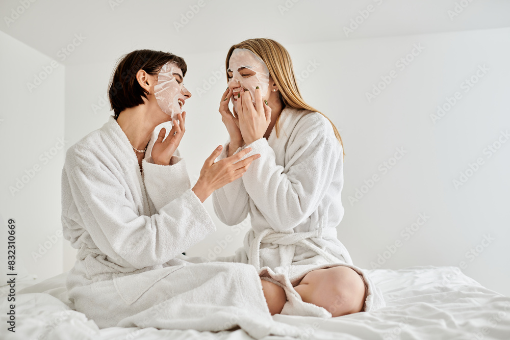 Two women in bath robes sitting on a bed, calmly wearing face masks for a spa-like moment of relaxation. Two women in bath robes sitting on a bed, calmly wearing face masks for a spa-like moment of relaxation.