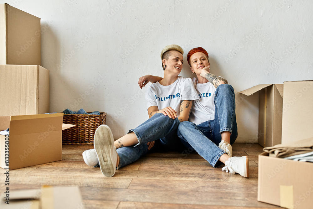 A young lesbian couple in volunteer t-shirts sit together on the floor, engaged in charity work. A young lesbian couple in volunteer t-shirts sit together on the floor, engaged in charity work.