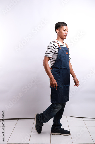 Full body side view smiling happy young asian man barista barman employee wear blue apron work in coffee shop walking going isolated on white background. Small business startup concept photo