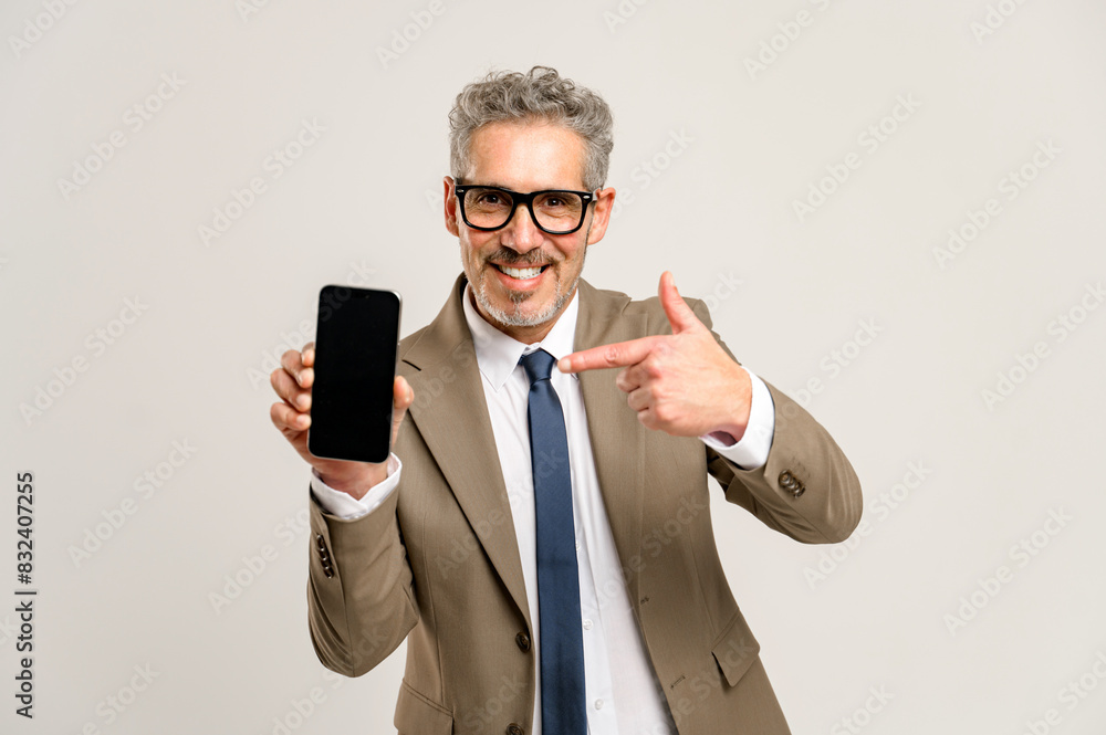 With a charismatic smile, a grey-haired businessman points to a smartphone with an empty screen, suggesting a place for advertising, copy space. Man in suit paying attention on pone display, mock up With a charismatic smile, a grey-haired businessman points to a smartphone with an empty screen, suggesting a place for advertising, copy space. Man in suit paying attention on pone display, mock up