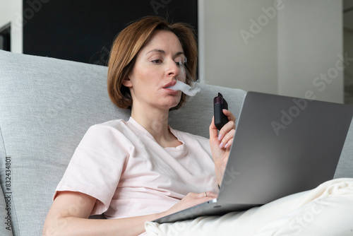 Young woman using disposable electronic cigarette during work on laptop at home 