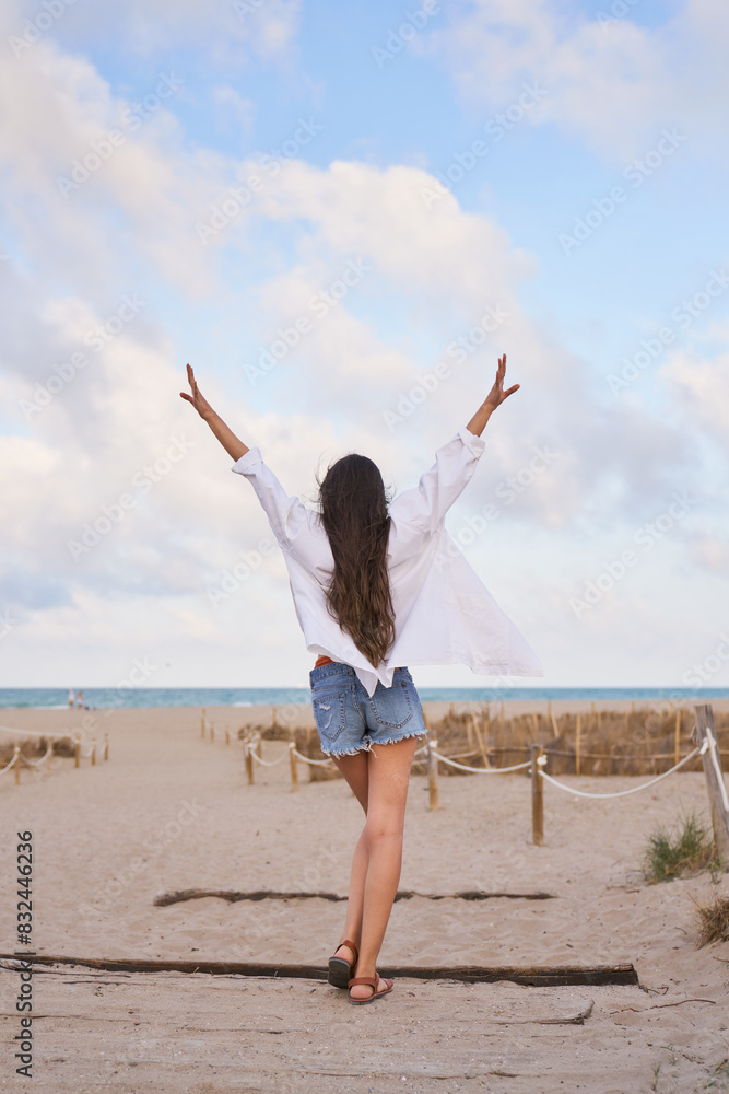 A woman stands on a beach with her arms raised in the air