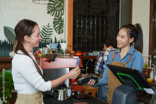 Fotografie Inside a cozy coffee shop, Asian female barista in denim and apron accepting a credit card from another female