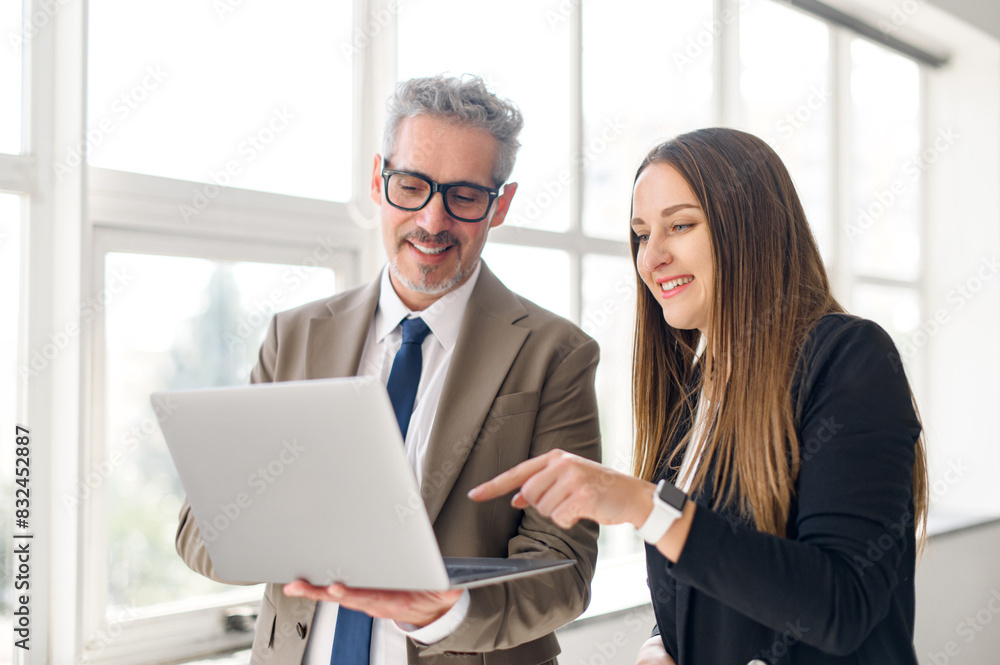 Young female professional points to screen of a laptop while a senior businessman with a jovial expression looks on, indicative of a collaborative and educational moment in a light-filled office space Young female professional points to screen of a laptop while a senior businessman with a jovial expression looks on, indicative of a collaborative and educational moment in a light-filled office space