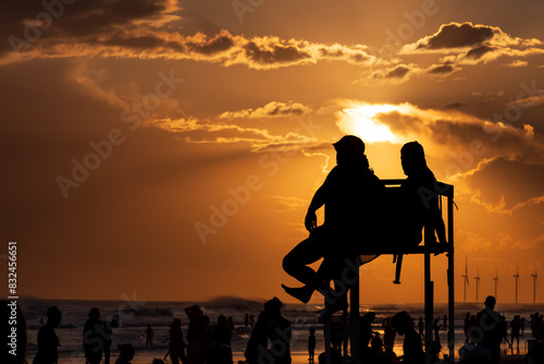 Silhouettes of lifeguards on an elevated bench and bathers on a beach at sunset.