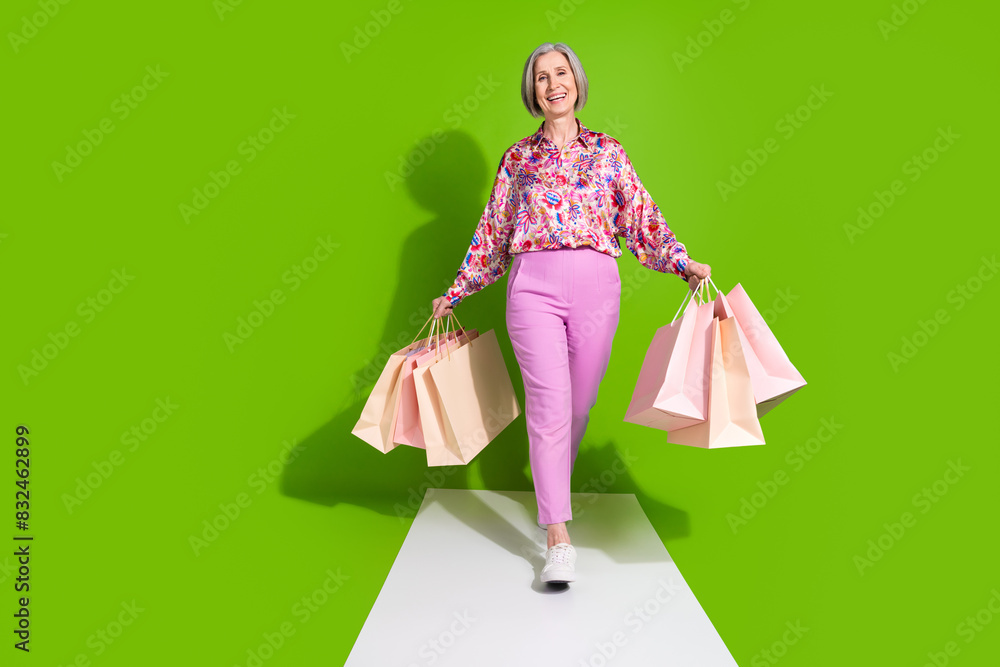 Full length photo of good mood pretty lady wear pink blouse walking way holding bargains empty space isolated green color background Full length photo of good mood pretty lady wear pink blouse walking way holding bargains empty space isolated green color background