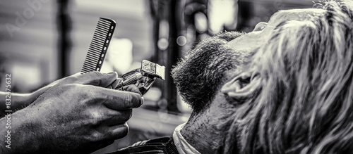 Close-up of man haircut, master does hair styling in barber shop. Hands of a hairdresser with a beard clipper. Barber works with a beard clipper. Hipster client getting haircut. Black and white photo