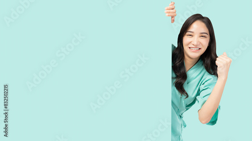 Confident young Asian female nurse peeking around a blank board, smiling and giving hand raised, wearing green medical scrubs. The image captures a positive and encouraging healthcare professional  