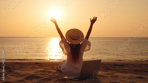 Work from anywhere. Rear view of young woman, female freelancer in straw hat working on laptop, keeping arms raised and cellebrating success while sitting on the tropical sandy beach at sunset  photo