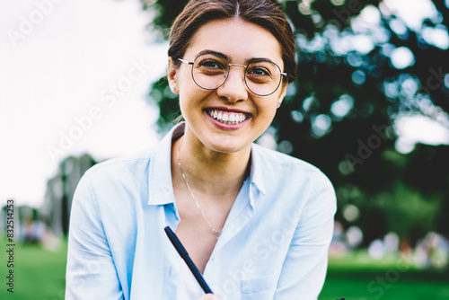 Portrait of cheerful young woman in spectacles enjoying learning outdoors sitting on grass in park, happy female student satisfies with good mark for project looking at camera resting on campus