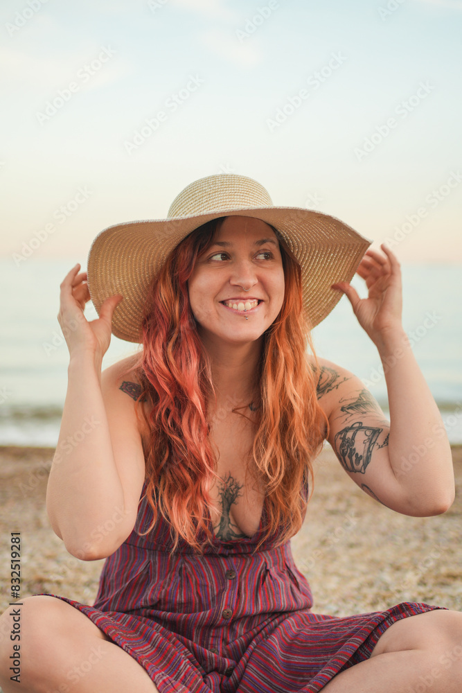 A woman with red hair and tattoos is sitting on the beach wearing a straw hat. She is smiling and she is enjoying her time