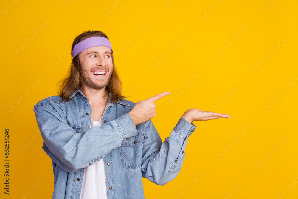 Photo portrait of attractive young hippie man point look palm hold empty space wear trendy denim outfit isolated on yellow color background