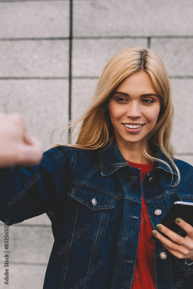 Young charming smiling blonde dressed in stylish jeans clothes downloading application from network using cellphone and internet connection during giving knock fists to male best friend outdoors Young charming smiling blonde dressed in stylish jeans clothes downloading application from network using cellphone and internet connection during giving knock fists to male best friend outdoors