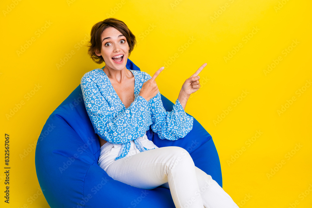 Portrait of astonished girl dressed print blouse sit on pouf indicating at impressive sale empty space isolated on yellow color background Portrait of astonished girl dressed print blouse sit on pouf indicating at impressive sale empty space isolated on yellow color background