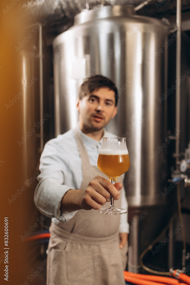Happy brewer. Happy young male brewer in apron holding glass with beer and looking at it with smile while standing in front of metal containers Happy brewer. Happy young male brewer in apron holding glass with beer and looking at it with smile while standing in front of metal containers