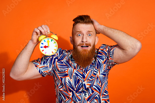 Photo of impressed shocked guy dressed print shirt arm head showing clock isolated orange color background