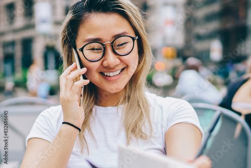 Cheerful asian girl enjoying communication with friend discussing plans while sitting in cafe in downtown,happy female student using connection in roaming for making calls abroad with cheap tariffs