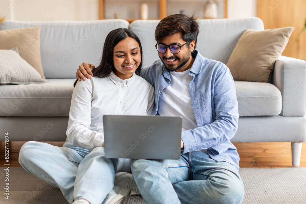 Indian man and woman are seated on the floor, both focused on a laptop screen in front of them. They appear engaged in whatever is displayed on the computer, leaning towards each other. Indian man and woman are seated on the floor, both focused on a laptop screen in front of them. They appear engaged in whatever is displayed on the computer, leaning towards each other.