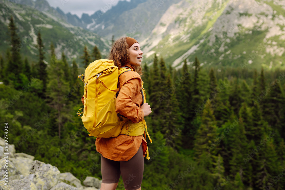 Naklejka premium A smiling female traveler with a yellow hiking backpack enjoys the beautiful scenery of the majestic mountains. Travel, adventure. Concept of an active lifestyle.