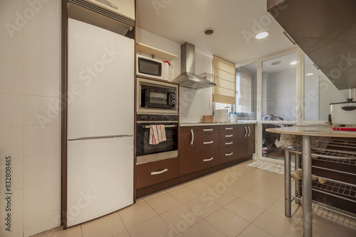 A kitchen with brown wooden furniture with a glazed terrace in the background and a column with integrated appliances