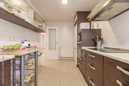 A kitchen with brown wooden furniture, a column with integrated appliances, a light countertop and cream-colored stoneware floors