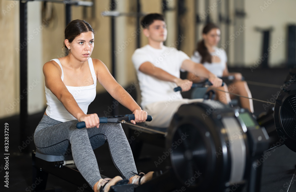 Workout girl cross training exercising cardio using rowing machine with group of people in crossFit club