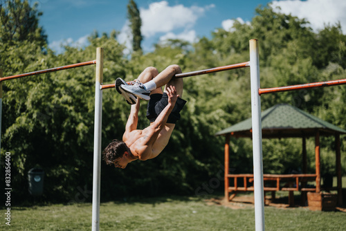Man doing calisthenics on a horizontal bar in an urban park with lush greenery and a gazebo on a sunny day.