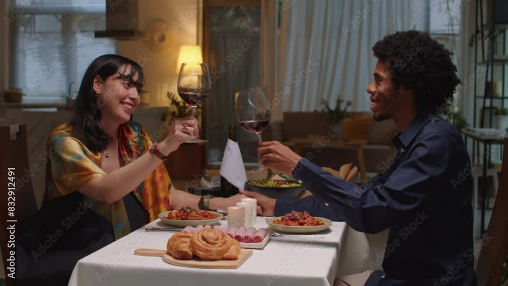 Side view of young affectionate couple toasting with glasses of red wine while sitting by festive table at Valentines Day Side view of young affectionate couple toasting with glasses of red wine while sitting by festive table at Valentines Day