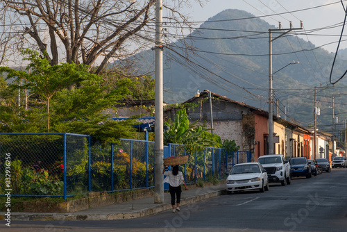 Fototapeta Naklejka Na Ścianę i Meble -  Young local woman carrying a large straw basket on top of her head on a small narrow street of a rural town, out of focus mountain landscape in the background. Ahuachapan, El Salvador.