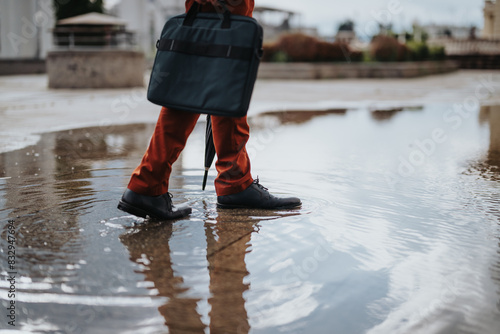 Close-up of businessperson with briefcase walking through a puddle reflecting city buildings after rain. Concept of urban life, work commute, and weather challenges.