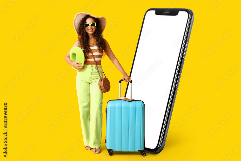 Young African-American woman with suitcase, pillow and big mobile phone on yellow background