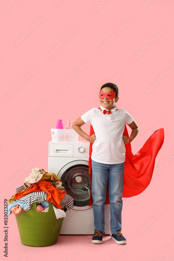 Little African-American boy in superhero costume near washing machine and basket with laundry on pink background
