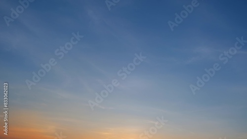 A tranquil sunset with a clear blue sky gently transitioning to warm hues of orange and yellow near the horizon. Delicate wispy clouds are scattered across the sky. Sunset sky background. 