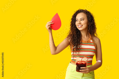 Young female African-American donor with applied medical patch, paper blood drop and pack for transfusion on yellow background photo