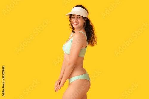 Beautiful young African-American woman in stylish turquoise swimsuit and cap on yellow background
