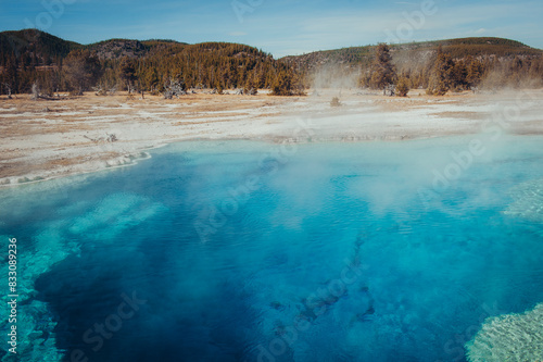 Sapphire blue geothermal pool amidst steaming hot springs on a bright day. © NILSEN Studio